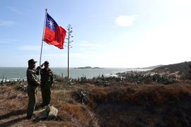 Two veterans raising a Taiwan flag on Kinmen. The sea is behind them and the landscape is rocky.