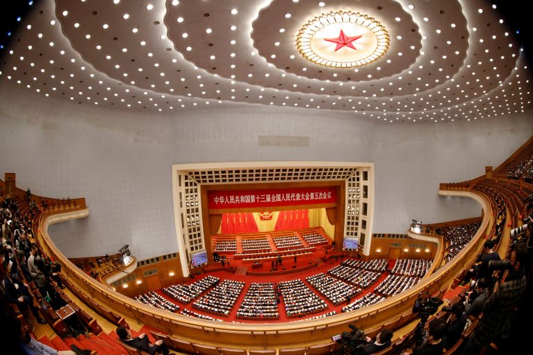 A wide-angle view of the NPC at the Great Hall of the People in Beijing. There are balconies of seating. The stage below has rows of seats for officials and a red backdrop with a yellow hammer and sickle. There is a red star in the light at the centre of the roof