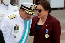 Honduras President Xiomara Castro talking to the Chairman of the Joint Chiefs of Staff at a military event. He is in ceremonial uniform and stooping slightly to hear her. She is wearing a burgundy jacket and sunglasses.