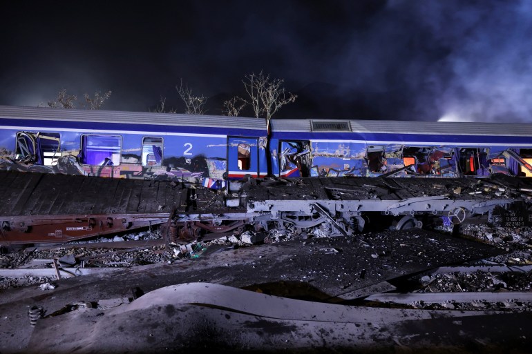 Two damaged train carriages following the collision in Greece. The doors have been pushed in and windows broken or pushed out. There are transporters and debris from the cargo train in front. Clouds of smoke are rising behind in the night sky