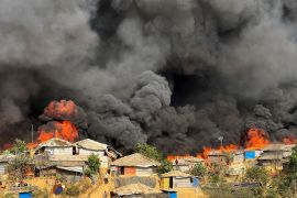 Clouds of black smoke filling the sky in the Balukhali refugee camp. There are orange flames consuming the wooden and bamboo huts