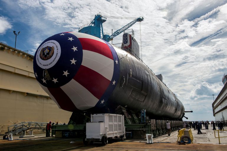 A Virginia-class submarine on the dry dock at a shipbuilding yard. Its nose is decorated in the colours of the United States. There is a crane behind.