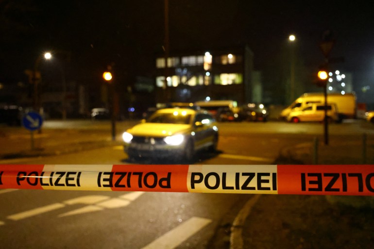 Red and white police tape with the upside down and upright word POLIZEI running along the tape. There is a car behind the tape with its lights on. It is nigh and everything is bathed in the warm yellow light of steep lamps.