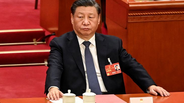 Xi Jinping sitting at his desk at the NPC. There are papers on the table and a tea cup. He has both arms reaching out to touch the desk.