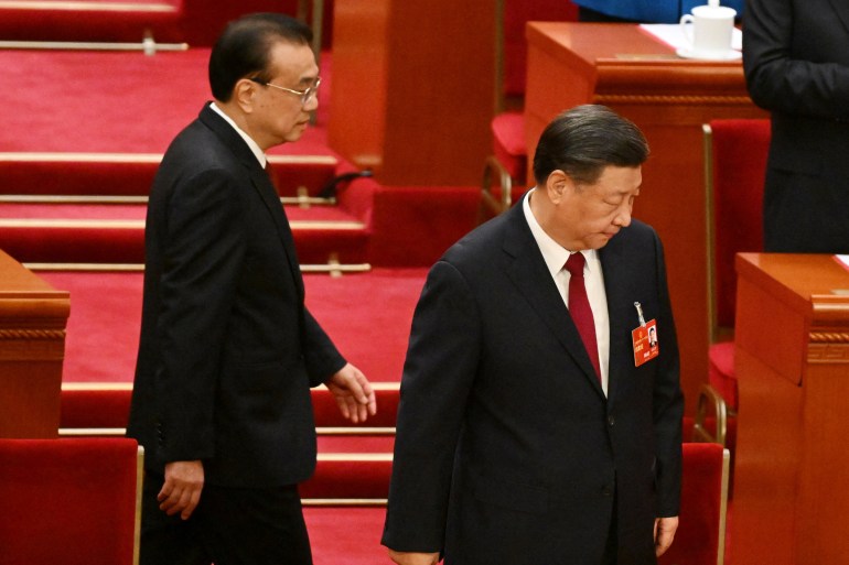 Xi Jinping taking his seat as ex Premier Li Keqiang walks behind him.