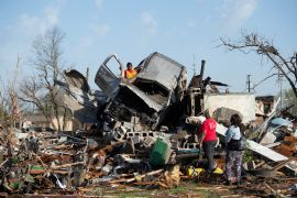 Residents of Rolling Fork, Mississippi, survey damage from a tornado. A visibly-damaged car, possibly a SUV, is lying on top of a mound of debris.