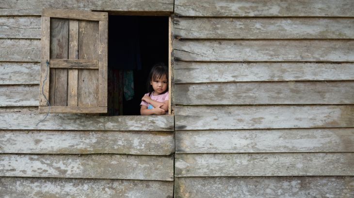 A young girl looks from the window of a wooden house. She is wearing a pink t-shirt and has her arms around her chest. She looks quite serious.