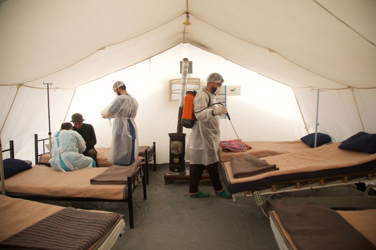 Inside a tent that has two rows of beds on each side, their tails facing each other, with a walkway in between. There is a heater at the end of the walkway, at the back of the tent. Three medical workers, wearing personal protective equipment are also near the back. One has a tank strapped to his back and is spraying an empty bed. Another two are at the back of the tent, taking notes from a patient sitting on a bed.