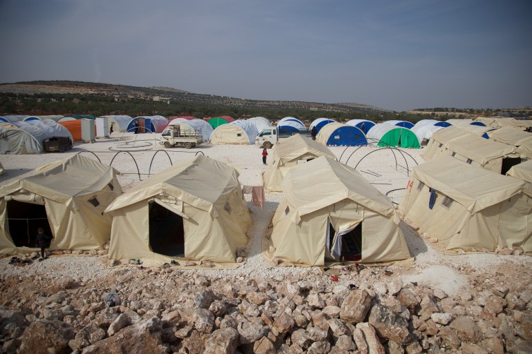 A view of tents set up in a sparse, desert-like area. Some at the front are beige while at the back are multi-coloured tents.
