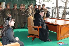 Kim Jong Un seated at a wooden desk in a tent-like shelter to watch a missile test. He is surrounded bt military officials in uniform. At the front of the photograph his young daughter is sitting on a chair.