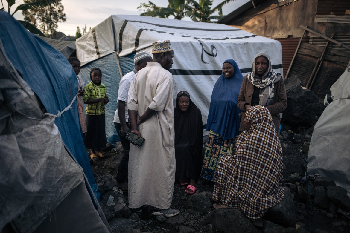 The imam of the mosque of the informal camp talks with other displaced people before the breaking of the Ramadan fast. The scene is poorly lit as it is almost nightfall. A half-dozen people are listening, smiling and talking with the imam. Most are standing with two people sitting in front of the religious leader.
