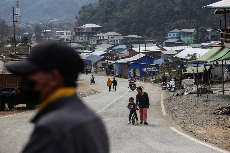 A street in a refugee camp at the Myanmar-India border. A road is winding through it. There is a woman walking towards the camera with a child on a small bicycle. Other people are walking or on mopeds.