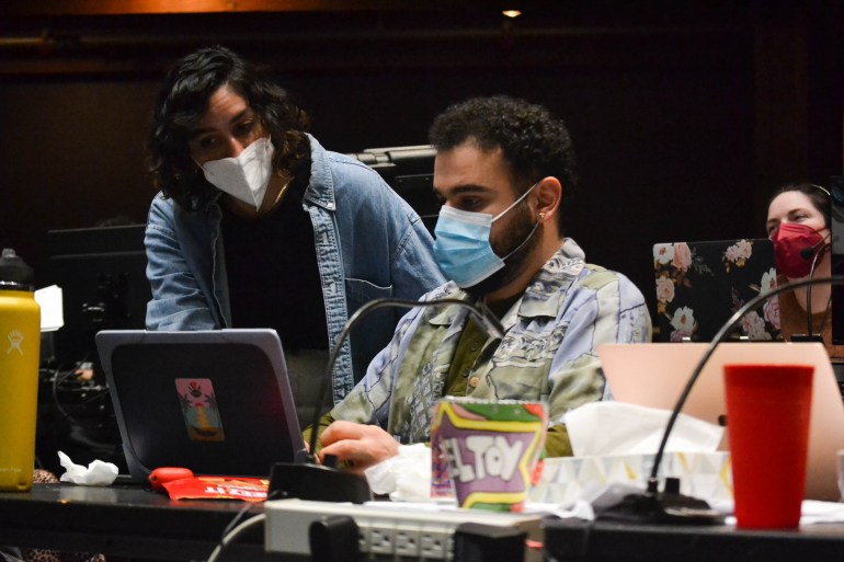 A man in a face mask sits at a laptop at a tech table, while a woman, standing, leans over to look at the computer