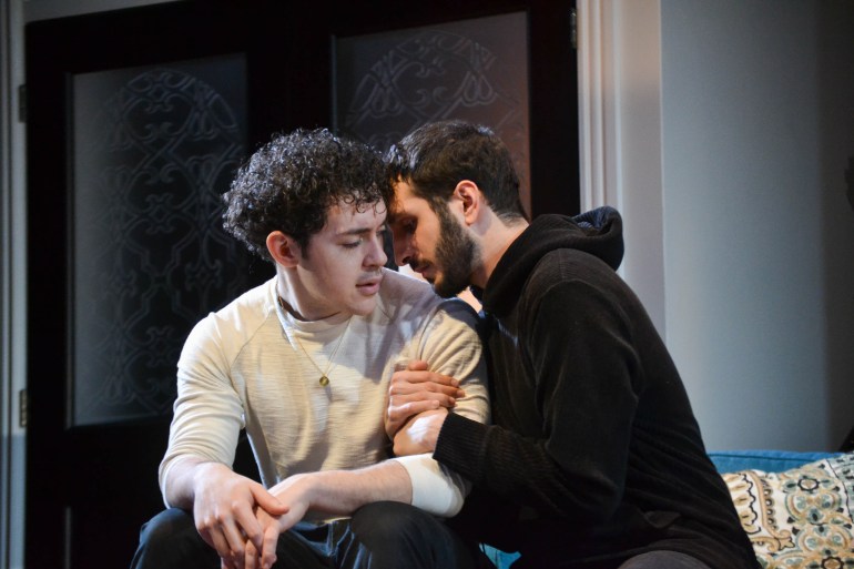 One actor in a black hoodie grabs the arm and leans his head against another actor as they perform a scene