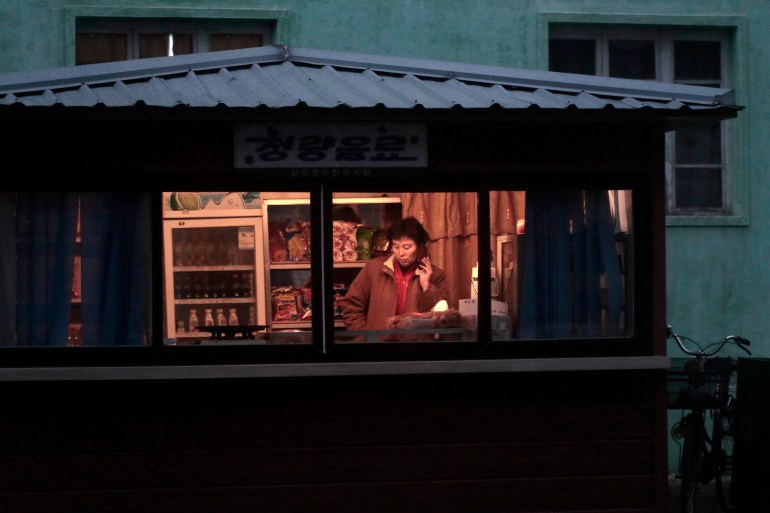 It seems to be nighttime but the rectangle of light behind the kiosk window shows a lady in brown jacket speaking on the phone. There is a fridge of drinks behind her.