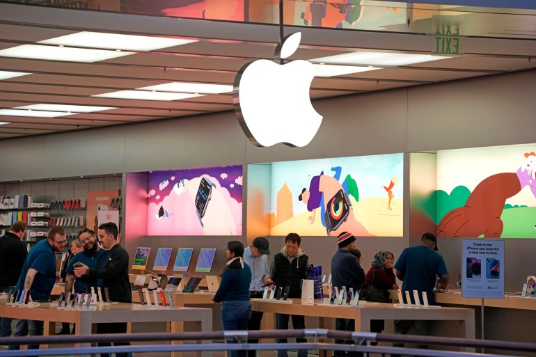 Looking into an Apple store, with bright displays, wooden tables with devices and an Apple logo hanging overhead