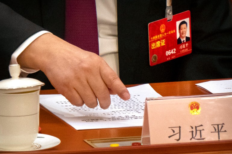 A close up of Xi Jinping's desk at China's National People's Congress showing him reaching out his finger to vote. There are papers on his desk and his red identity card is attached to his suit.