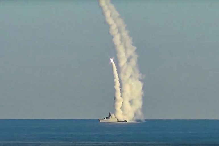 Long-range Kalibr cruise missiles are launched by a Russian military ship from an unknown location. Long trails of smoke can be seen tracing from the ship in the ocean and one flash of orange light, seemingly a missile being fired, can be seen in the middle of the screen.