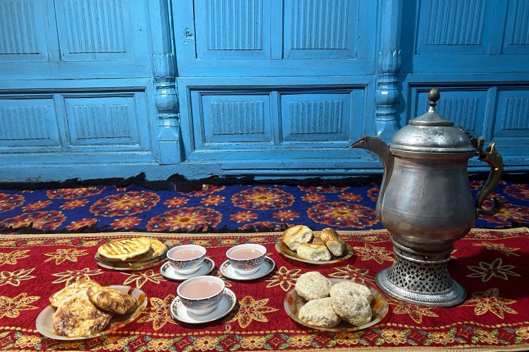 A samovar, three cups of noon chai and four plates of snacks laid out on a colourful carpet in front of a turqoise wooden wall