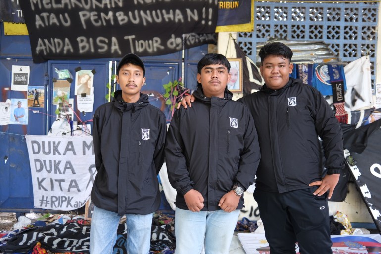 Galih Wahyu Prakoso with his friends Farel Izha Mahendra (left) and Cheva Octatista (right), who are members of a local football fan club. They are wearing black jackets and standing outside the stadium where their friends were among 135 people who died. There are tributes to the dead behind them.