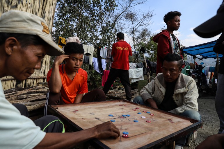 Chin refugees playing the board game carrom. There is a board between the four of them with coloured tokens in red blue and white. They look very intent. There are other people behind them, and some laundry on a washing line.