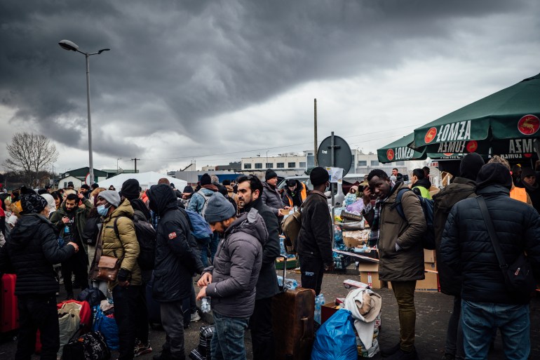 A photo of the border crossing at Medyka in Poland with lots of people standing around with suitcases with a ship in the background and a couple of umbrellas.