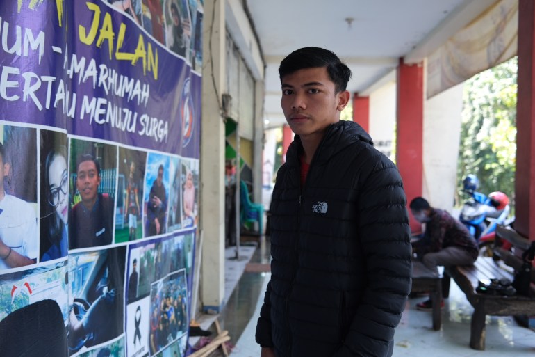 Nanda Rizky Kurnia Sandi outside the stadium. He is wearing a black puffa jacket. He is beside a banner paying tribute to the fans who died at the stadium.