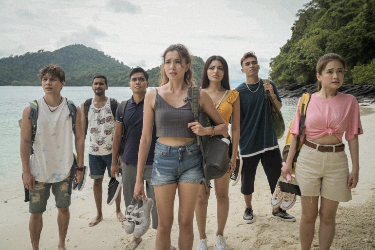 A still from the film Pulau, showing a group of young women and men gathered on the beach. They are in T-shirts and shorts