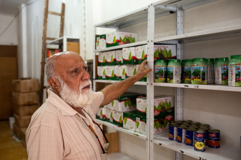 Shop owner Omar Dabage, 73, showcases some of his Arab products. He has a white beard and is holding onto a column of a shelf. Some shelves are bare, another has green jars on them, another some cans and three shelves behind him are stocked with boxes, all of the same design.