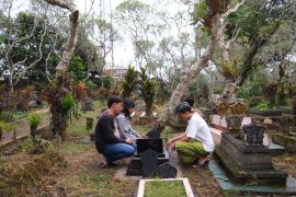 Wiyanto, his daughter and Septian’s friends pray together by his grave, which is near their family home. They are kneeling down. Wiyanto is on the left of the grave. There are gnarled trees around the cemetery.