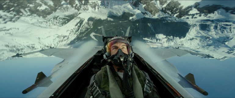 A scene from Top Gun: Maverick, showing Tom Cruise in the cockpit of a jet, upside down over a snowy mountain range.
