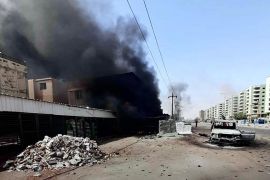 A battle-damaged street in Khartoum, Sudan, with black smoke billowing out of a building.