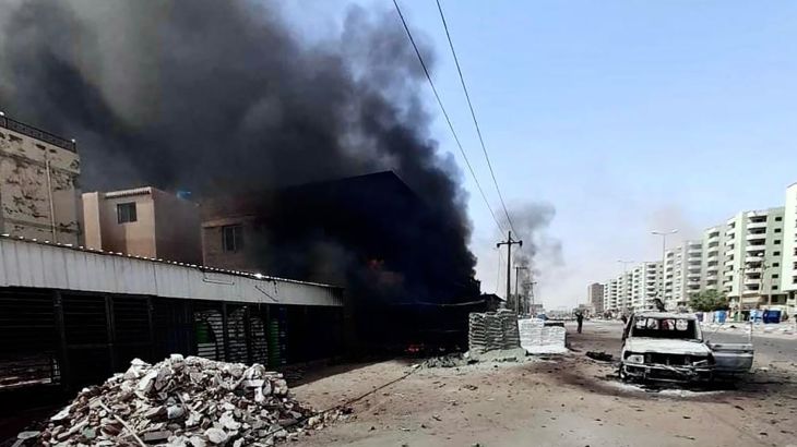 A battle-damaged street in Khartoum, Sudan, with black smoke billowing out of a building.