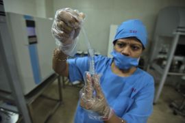 An embryologist at a fertility centre in India preparing a sample. She is wearing plastic gloves, a blue hair cover and mask with IVF written in red on the front.