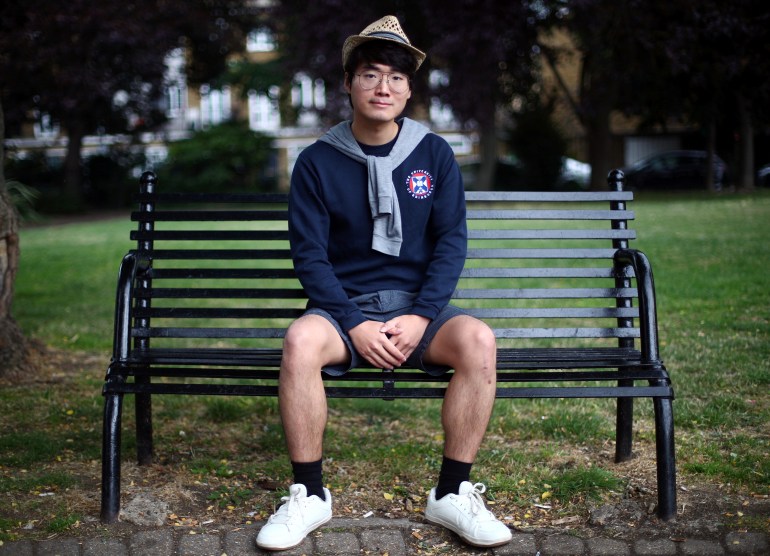 Simon Cheng posing for a photo. He is sitting on a bench in a London park. He is wearing blue shorts and a sweatshirt and a hat.