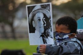 A little boy, bundled up in a face mask and jacket, lifts a sign with a black-and-white photo printed on it, depicting Emmett Till