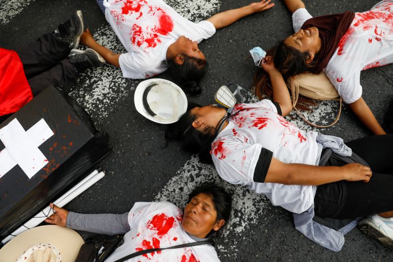 Protesters pretend to be dead on the concrete, wearing red-splattered white shirts. A fake coffin — black with a white cross — sits next to them.