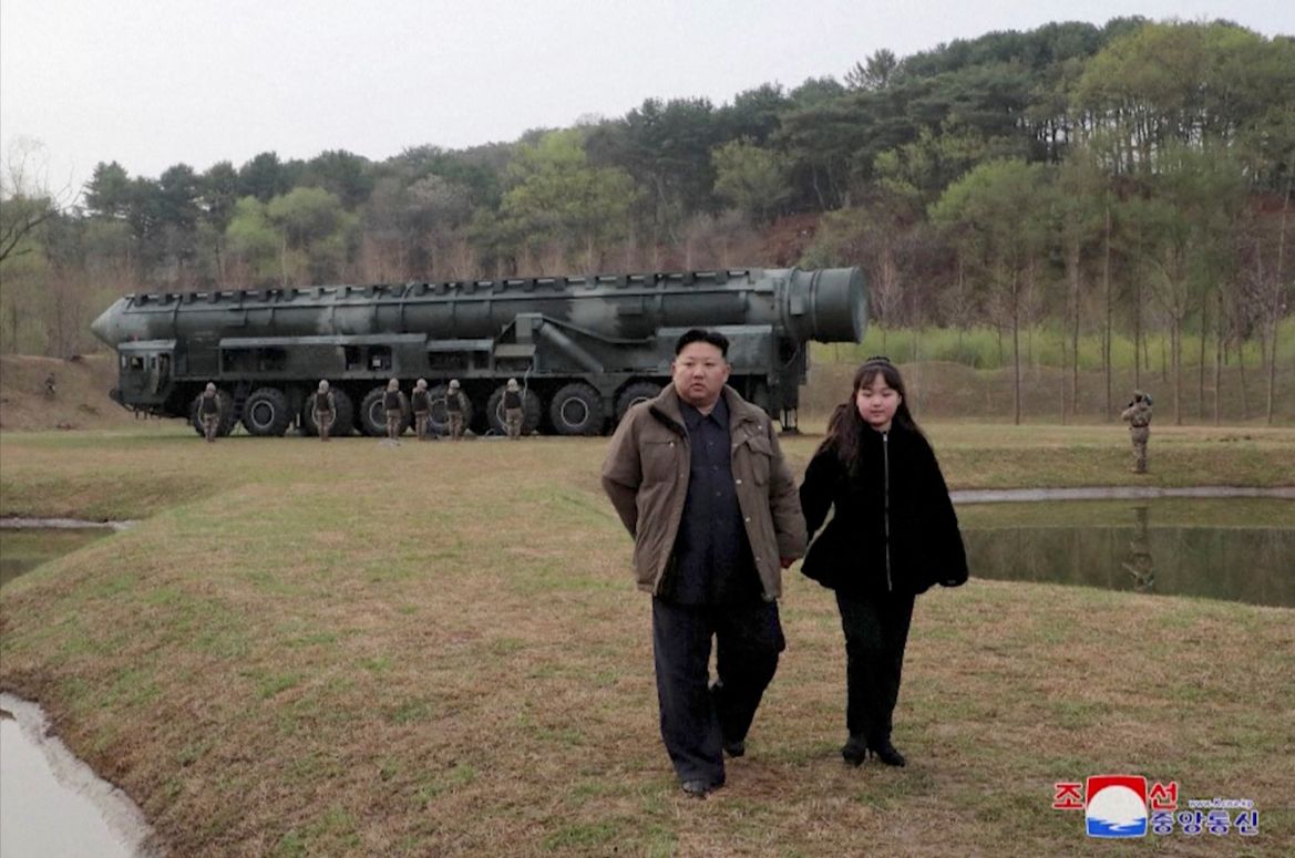 Kim Jong Un and his daughter Kim Ju Ae walking away from the missile and launcher towards the camera. She is wearing black while he's in black with a brown jacket.