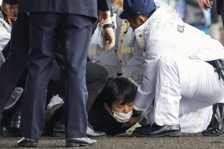 A man dressed in black and with a face mask lying face down on the ground. He is being held down by police officers but is looking up