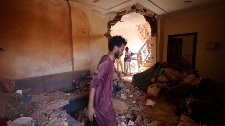 A man stands amid rubble in a Khartoum house hit during fighting between the army and RSF, The doorway behind hin is damaged, There is rubble all over the floor and one the large sofas against two walls