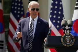 Biden, wearing sunglasses, answers a question next to a podium and in front of a row of US flags