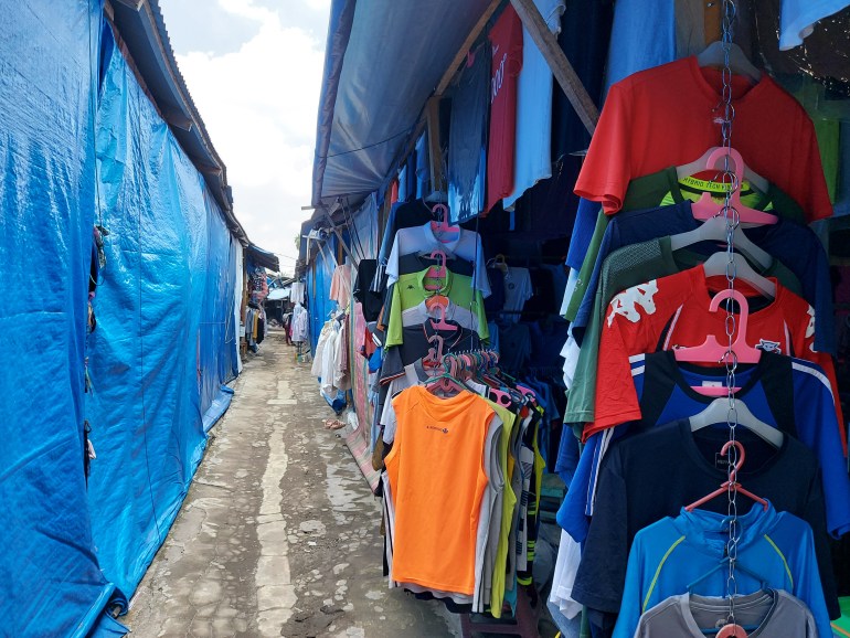 Colourful sport T-shirts and hoodies hang from racks in market stalls to the right of a walkway. On the left, the stalls are shut by a blue tarpaulin.