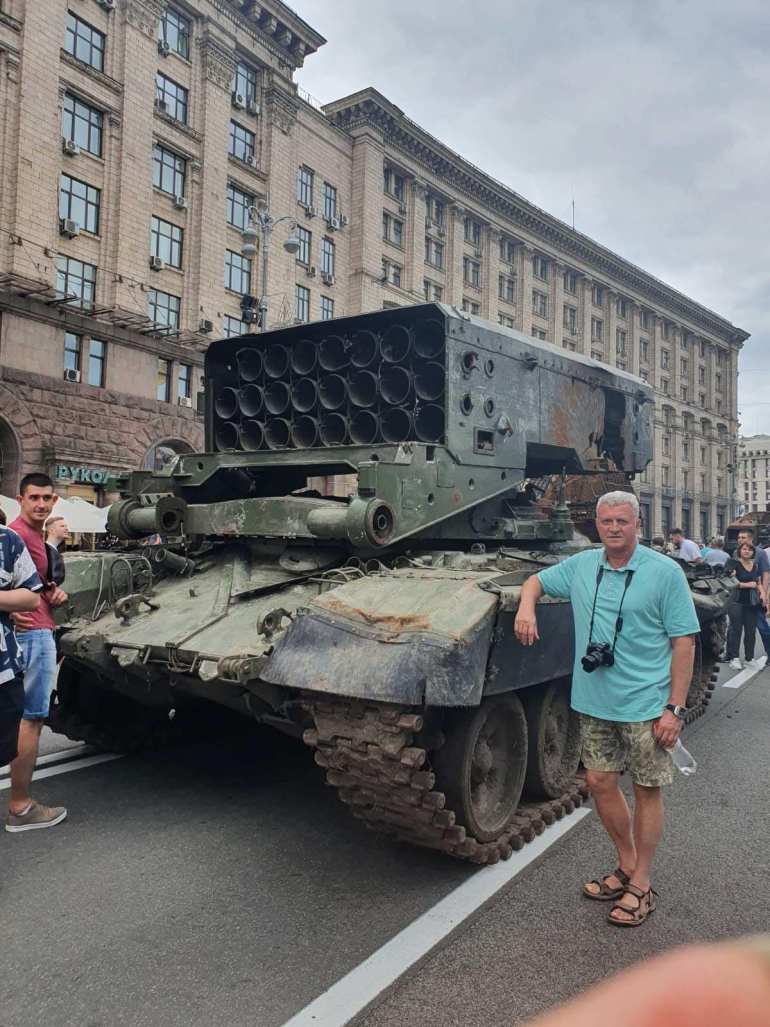 Oleksandr, wearing a blue T-shirt, shorts and with a camera around his neck, leans against a damaged military vehicle in the centre of Kyiv.