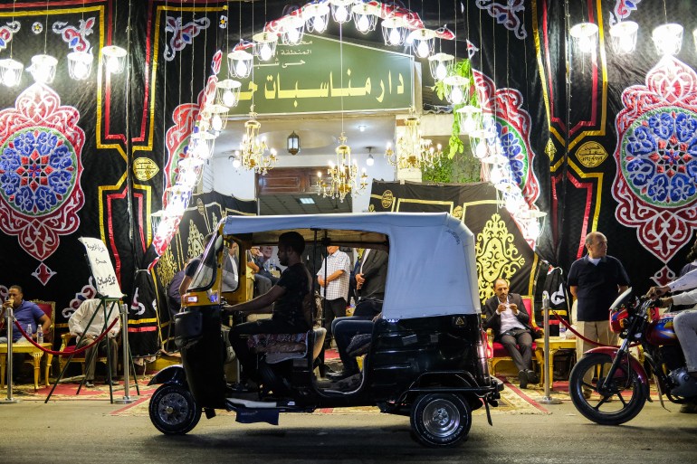 a tuk-tuk (motorised rickshaw) drives along a road past a funeral memorial service in the northern suburb of Shubra