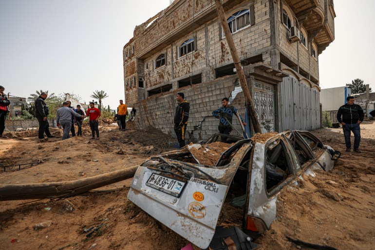 A destroyed car sits covered by sand, its roof caved in and back detached. People are milling around, surveying the damage. There is what looks to be an apartment block behind them that is intact but damaged.