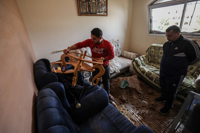 Maidi's son lifts a damaged side table from between a blue couch and a matching armchair. Maidi stands behind him with his arms folding behind his back. There is dirt on the rug on the floor and another two, mismatched couches.