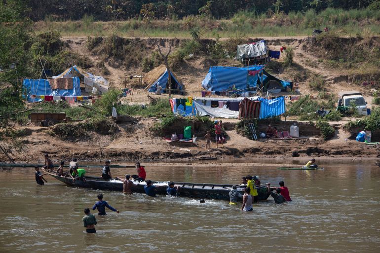 People taking food aid across the river to Myanmar villagers forced to flee by fighting between the military and rebel groups. There is an open boat crossing the river and tents made from blue tarpaulin on the opposite bank. Some people are wading in the river.