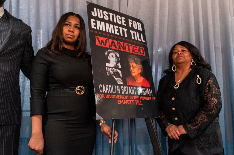 Two women stand on either side of a sign that reads: "Justice for Emmett Till. Wanted: Carolyn Bryant Donham"