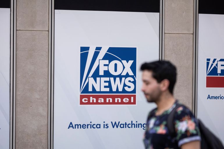 A man with a backpack walks by a Fox News logo, with the slogan "America is watching."