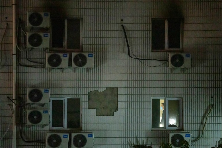An investigator inside one of the hospital rooms damaged in the fire. The photo is taken from outside and shows a series of windows with air conditioners below them and around. The room is lit up by the investigators torch. There are black smoke marks from some of the windows.